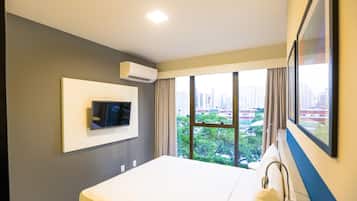 A hotel room with a bed, a mounted TV, an air conditioning unit, and a large window offering a city view.