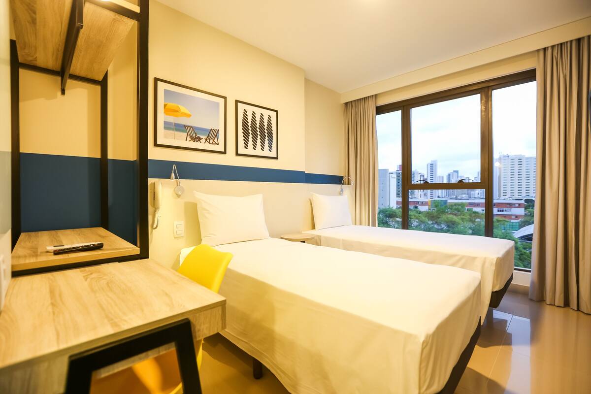 A hotel room with two beds, a desk, a chair, and a large window offering a city view.