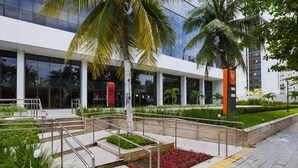 Un edificio moderno con fachada de vidrio, zona de estar exterior con barandillas de metal y un jardín ajardinado con palmeras.