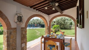 Ein überdachter Essbereich im Freien mit Tisch, gedeckt für das Frühstück, Steinwänden und Blick auf Pool und Garten.