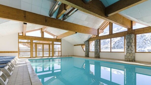 Une piscine intérieure avec des poutres en bois et des murs en pierre, entourée de fenêtres offrant une vue sur les montagnes enneigées.
