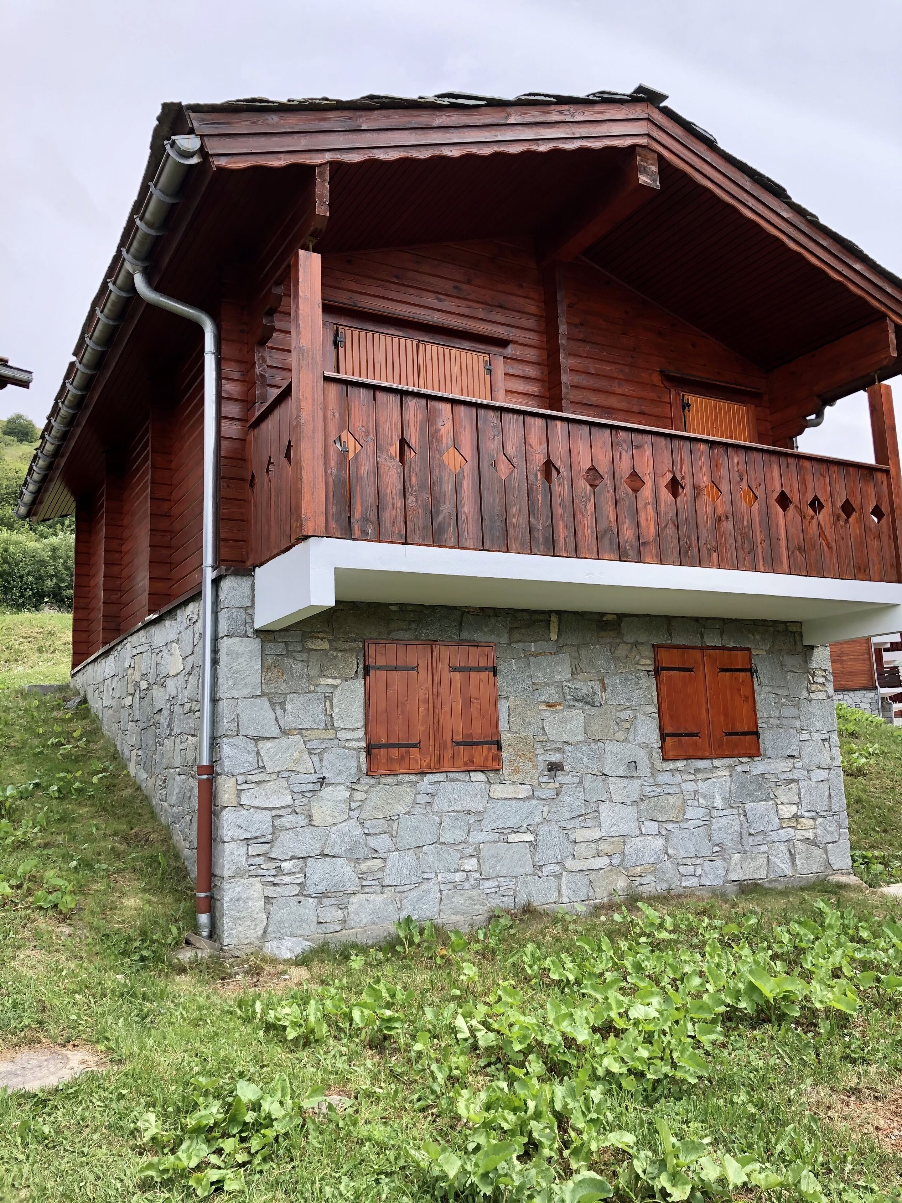A wooden chalet with a stone base, a balcony with wooden railings, and two wooden shutters.
