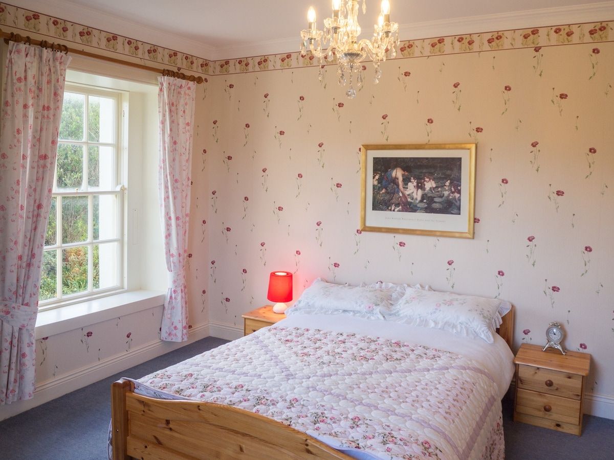 A bedroom with a wooden bed, a nightstand, a red lamp, a framed picture on the wall, and a window with floral curtains.