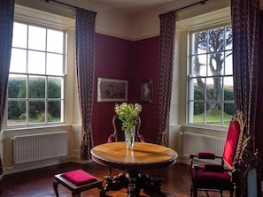 A room with a round wooden table, a red upholstered chair, and a vase with flowers. There are two windows with curtains, and a radiator is visible on the wall.