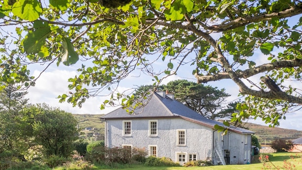 Ein Haus aus Stein mit einem Giebeldach, umgeben von einem grasbewachsen Feld und Bäumen.