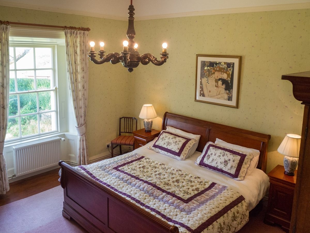 Un dormitorio con una cama de madera, un candelabro, una ventana con cortinas, una mesita de noche, una lámpara y un cuadro en la pared.