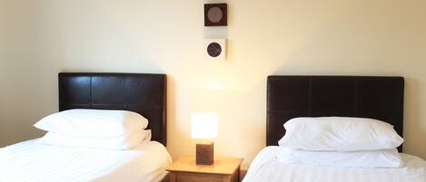 Une chambre d’hôtel avec deux lits, chacun doté de draps blancs et d’une tête de lit sombre. Une table de chevet se trouve entre les lits, avec une lampe et une petite horloge au-dessus.