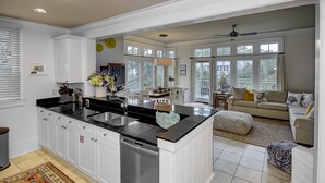 Cozinha moderna com armários brancos, bancada preta e lava-louças embutida. Há uma área de jantar com mesa e cadeiras, e uma sala de estar com sofá e ventilador de teto.