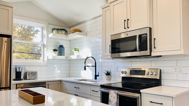 Una cocina moderna con armarios blancos, un refrigerador de acero inoxidable, un microondas, una estufa y un lavamanos. Hay plantas en macetas en los estantes y una ventana que proporciona luz natural.