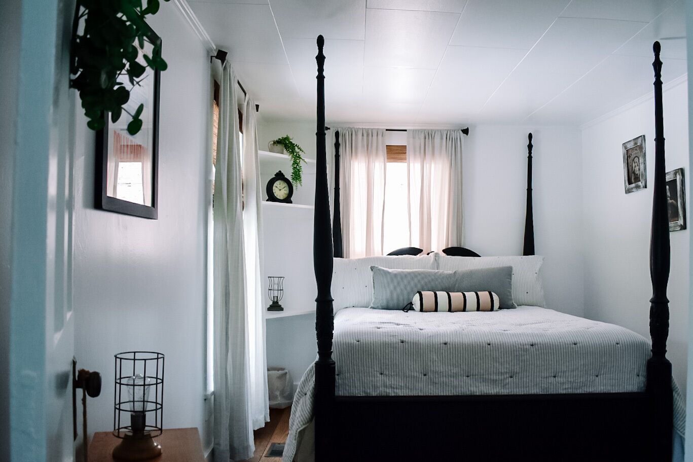 A four-poster bed with a striped bedspread and two pillows, a bedside table with a lamp, a clock, and a small plant on a shelf.