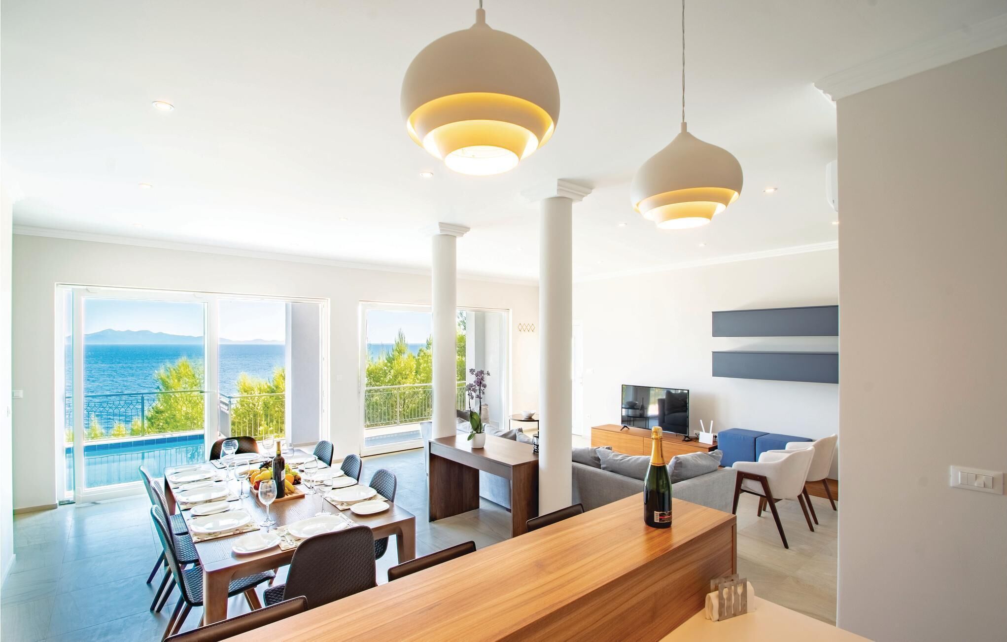 Ein großzügiger Wohnbereich mit Esstisch für sechs Personen, einer Couch und einem Fernseher. Große Fenster bieten Blick auf das Meer.
