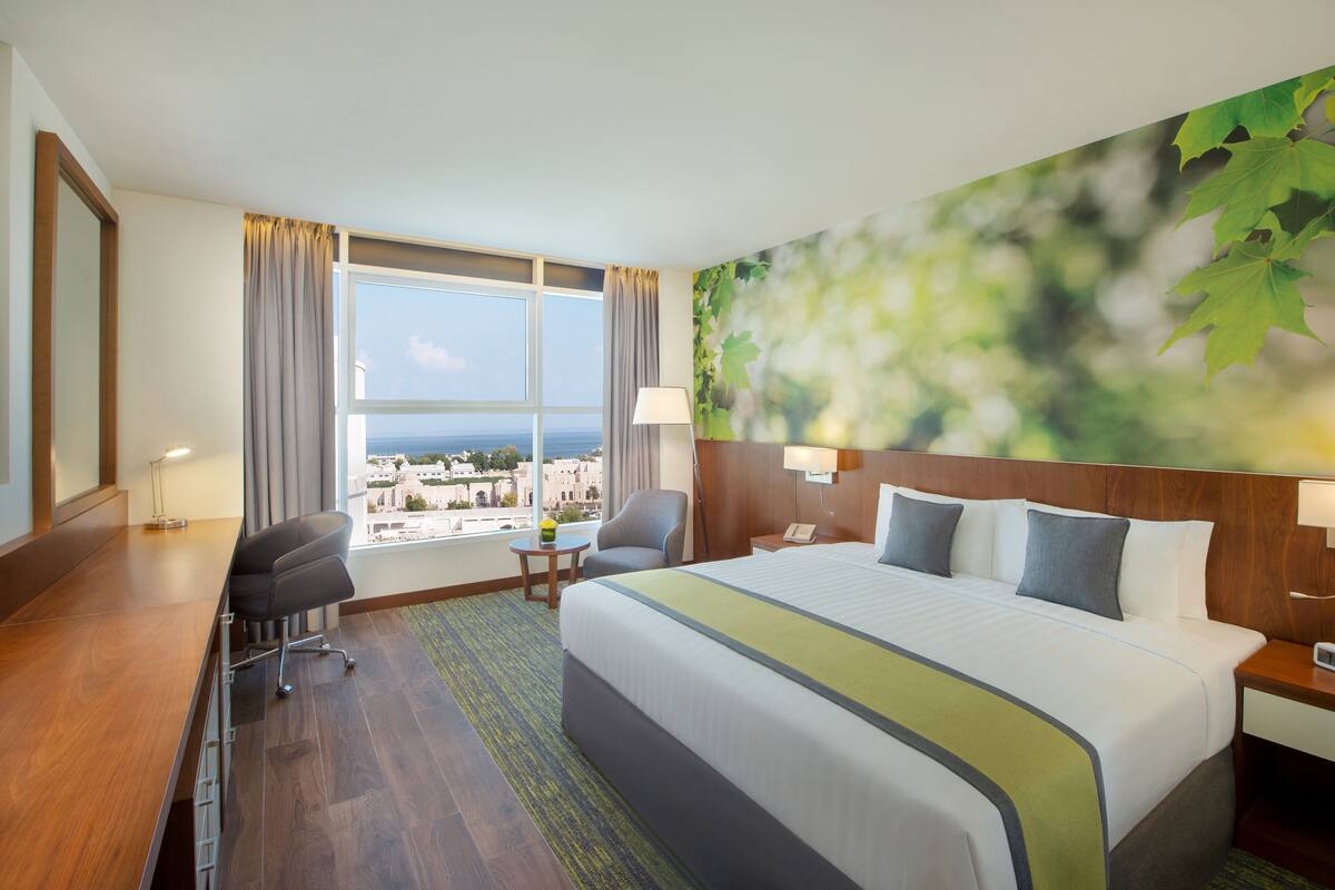 A hotel room with a large bed, a desk, a chair, a nightstand, and a view of the city and sea through the window.