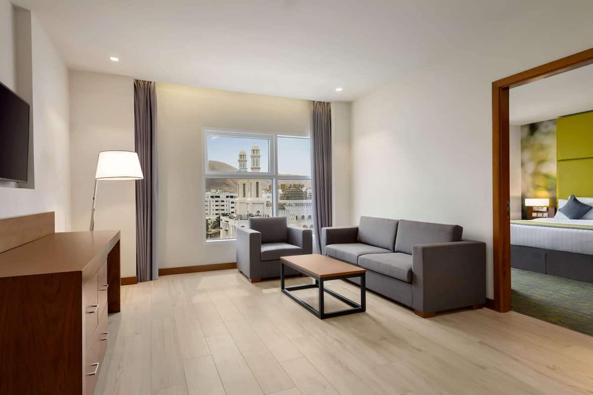 A modern hotel room with a sofa, armchairs, a coffee table, a TV, and a large window with a view of a mosque and buildings.