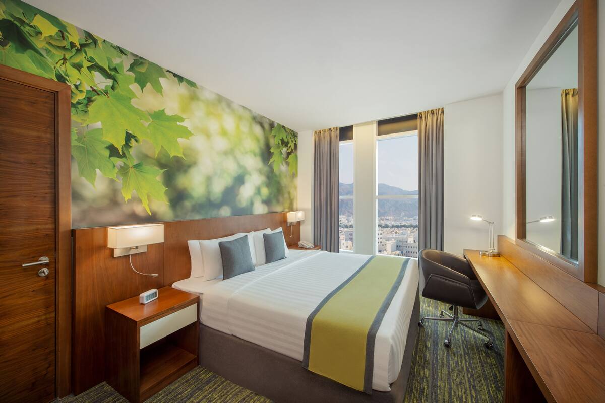 A hotel room with a large bed, a desk, a chair, a nightstand, a wall-mounted phone, and a window with curtains.