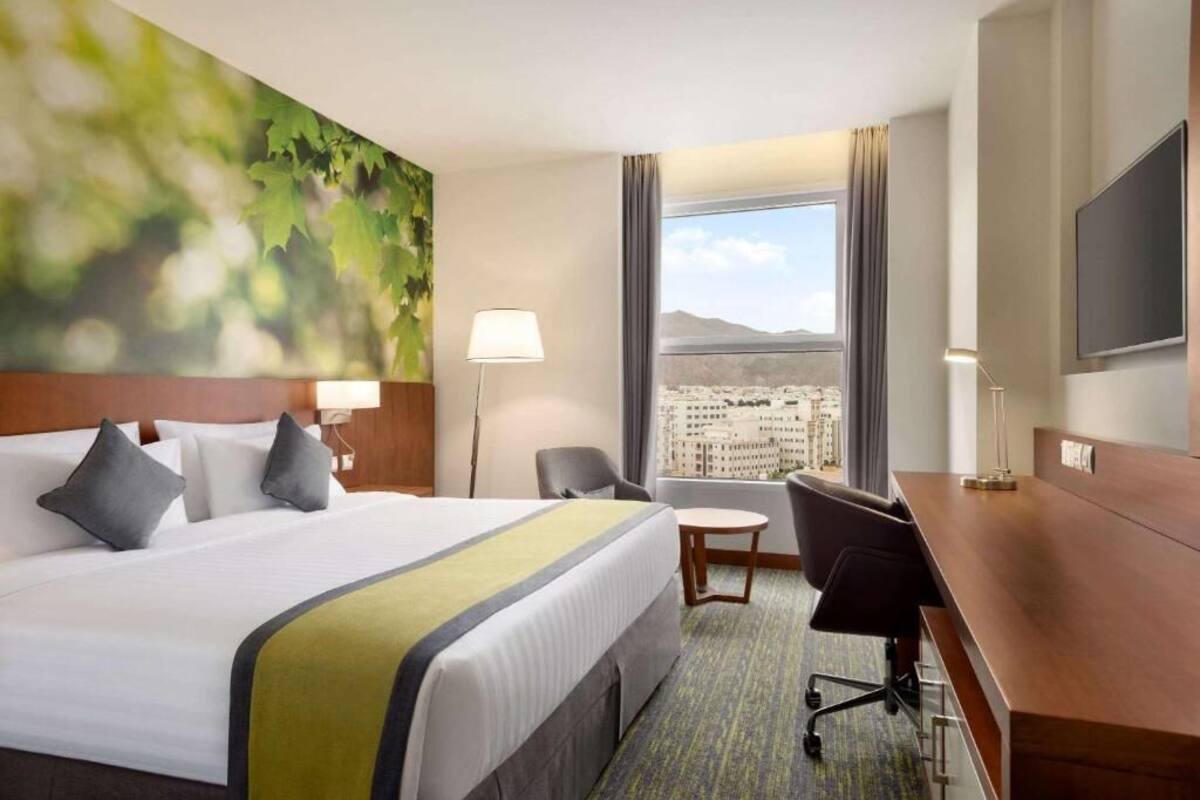 A hotel room with a large bed, a desk, a chair, a lamp, and a view of a cityscape through a window.