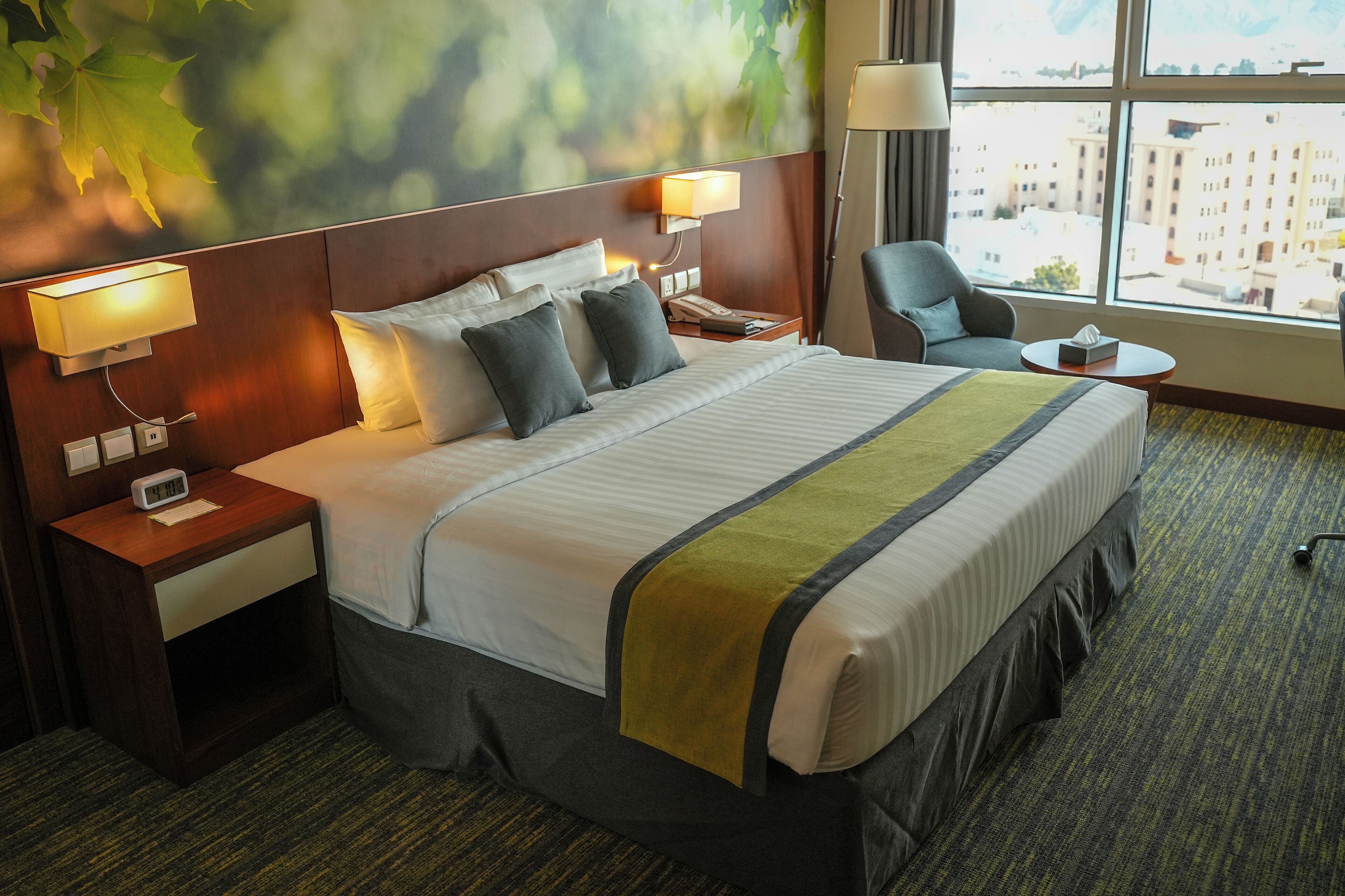 A hotel room with a large bed, two bedside tables, a chair, a small table, and a window with a city view.