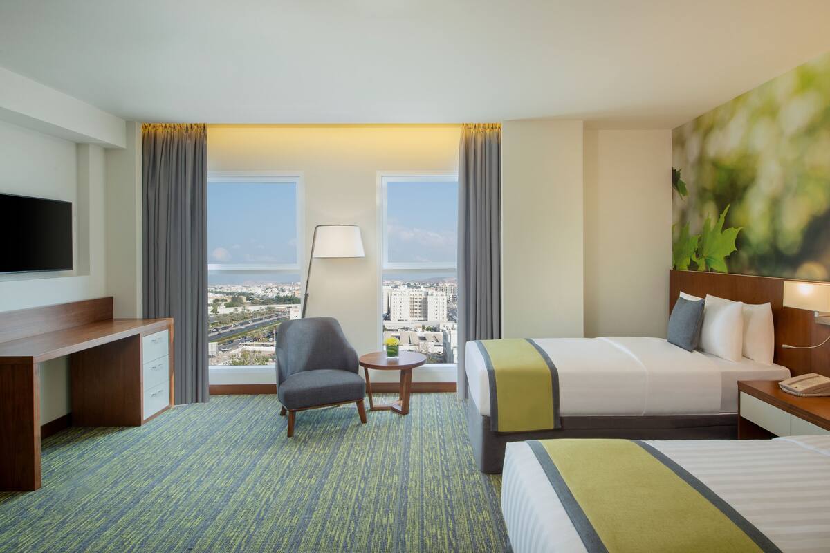 A hotel room with a bed, a desk, a chair, a TV, a lamp, and a view of the city.