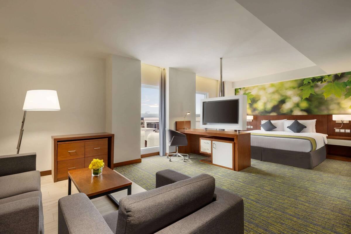 A hotel room with a bed, a desk, a TV, a sofa, a coffee table with flowers, and a view of the city.