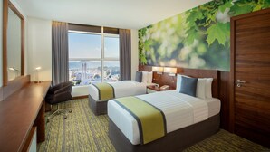 A hotel room with two beds, a desk, a chair, and a large window offering a city view.