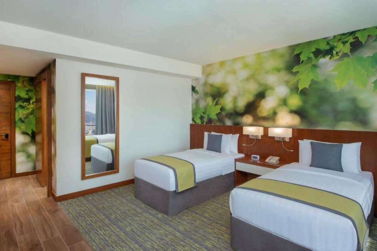 A hotel room with two beds, a mirror, a nightstand with a phone, and a view of a cityscape through a window.