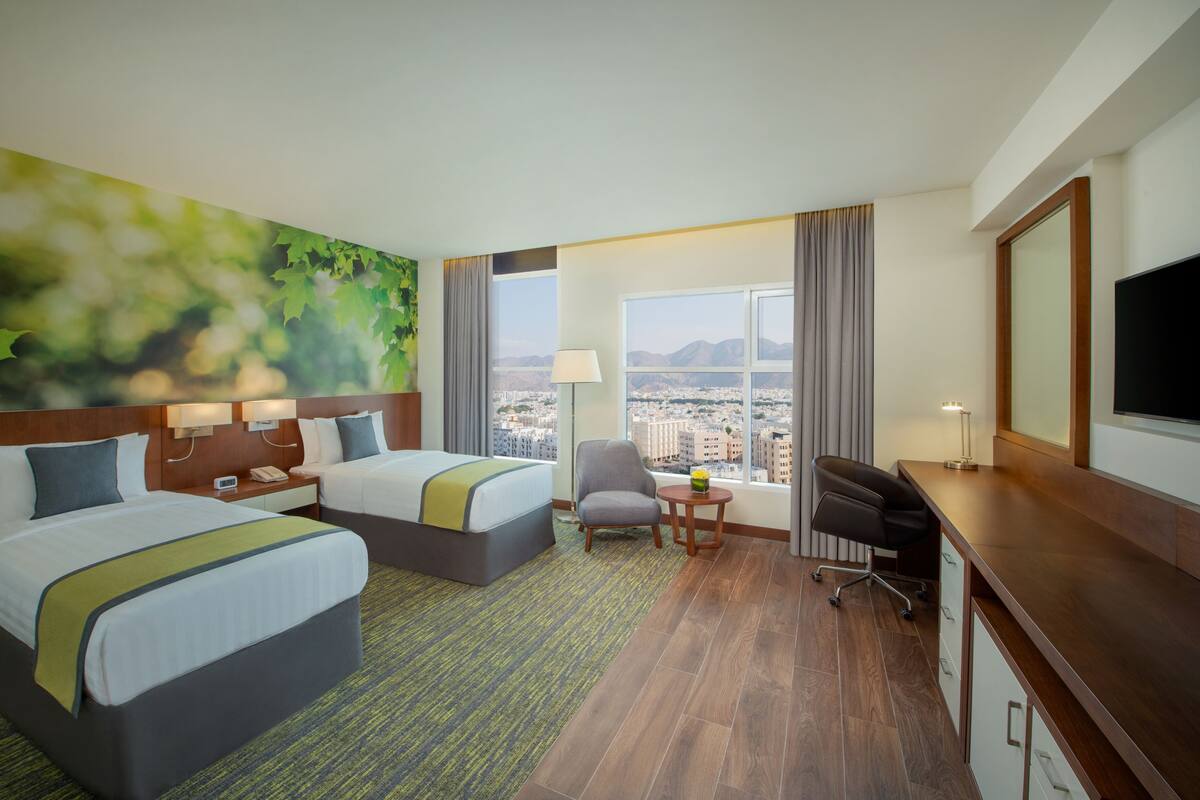 A hotel room with two beds, a desk, a chair, a TV, and a large window with a city view.