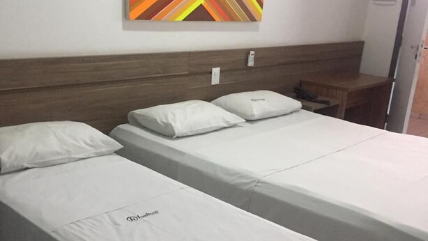A hotel room with two single beds, a wooden headboard, a small bedside table with a lamp, and a colorful abstract painting on the wall. - Samburá Hotel - Feira de Santana (Feira de Santana)