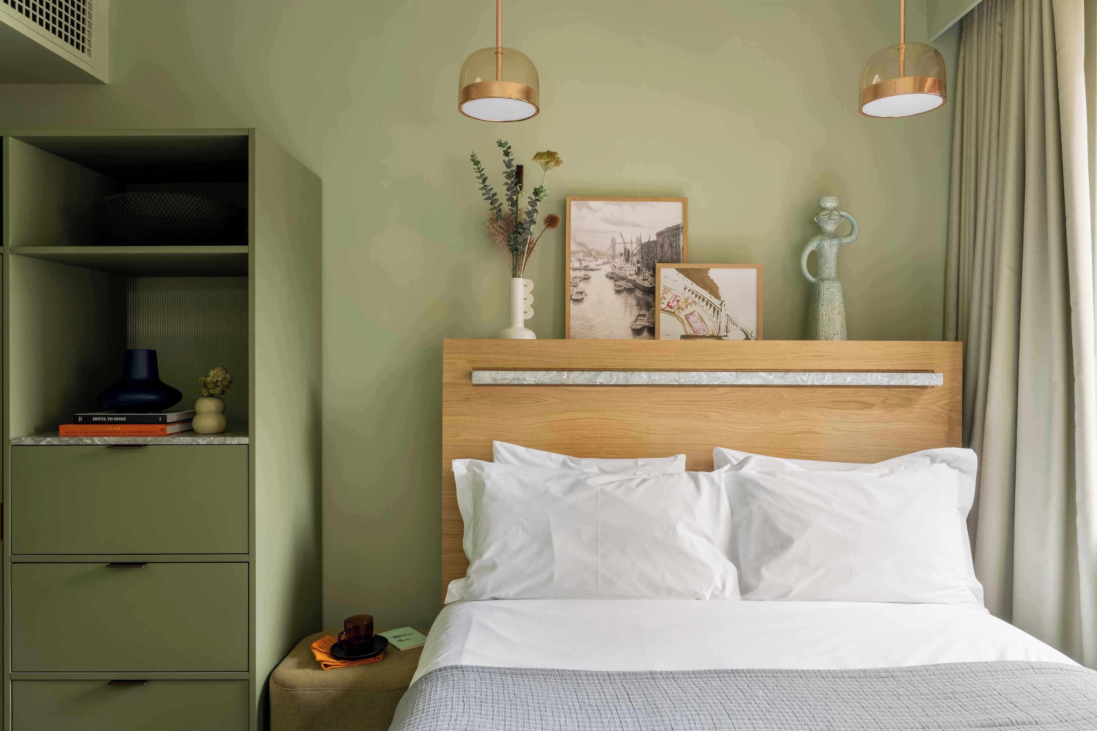 A neatly arranged bedroom with a wooden headboard, white bedding, a bedside table with a lamp, and a shelf with books and a vase.