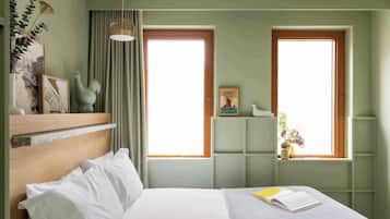 Un dormitorio con una cama, dos ventanas, un estante con adornos y una lámpara fijada a la pared.
