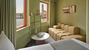 A hotel room with a bed, a small round table with a bowl, a sofa, a window with curtains, and a wall with framed pictures. - Locke At Broken Wharf (London)