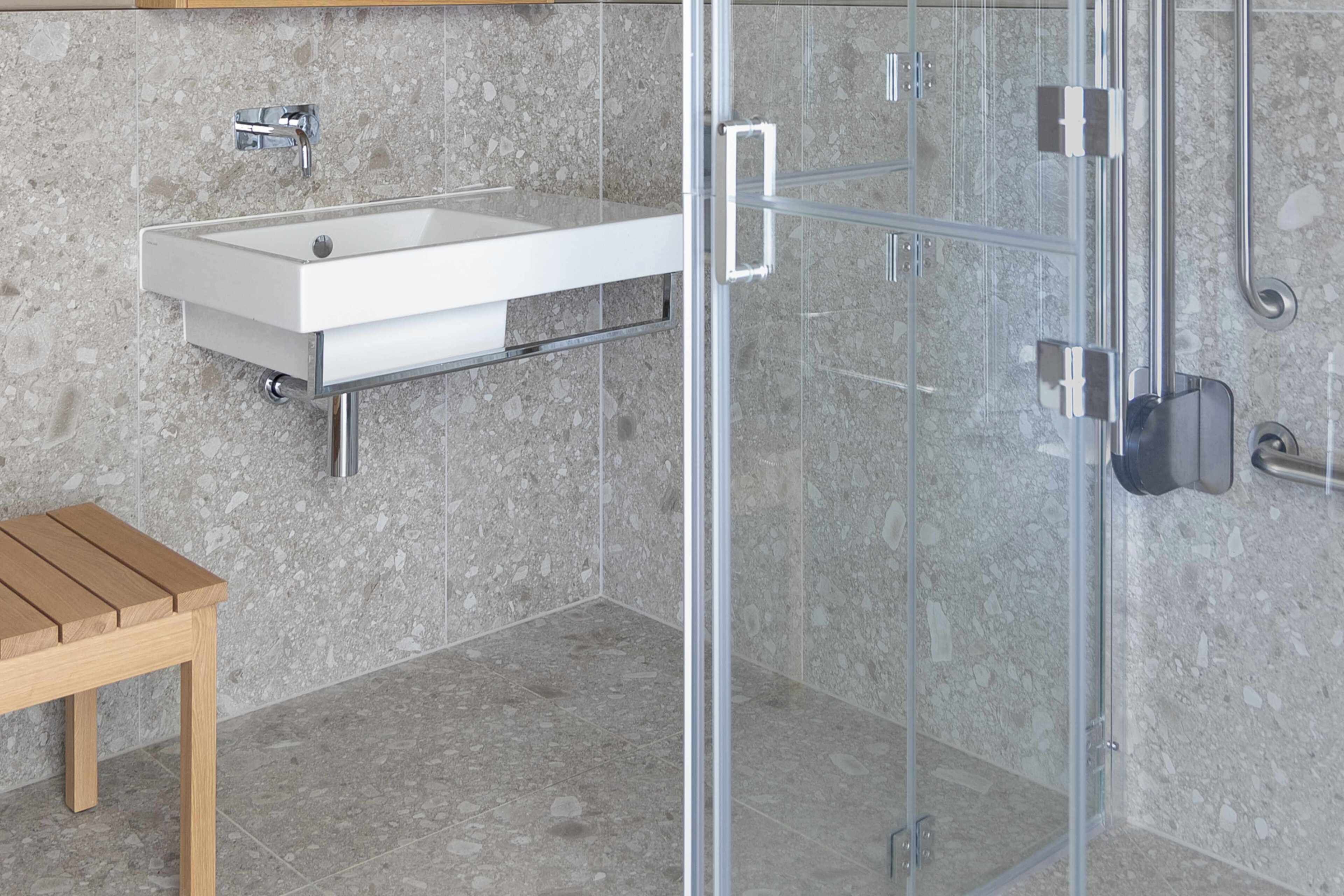 A bathroom with a glass shower enclosure, a white sink, and a wooden bench.