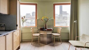 Een hoekappartement met twee ramen, een tafel met stoelen, een vaas met bloemen en uitzicht op de stad.
