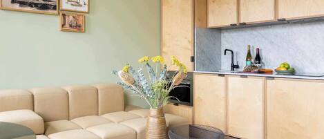 Una sala moderna con un sofá beige, una mesa de centro redonda con libros y un jarrón, y una zona de cocina con armarios de madera y encimera de mármol.