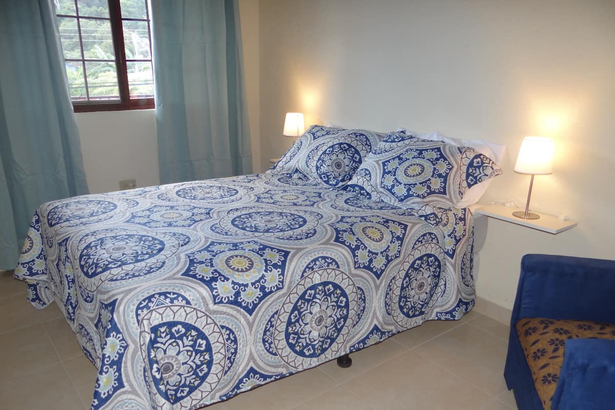 Un dormitorio con una cama cubierta por una colcha azul y blanca, dos lámparas de noche y una ventana con cortinas.