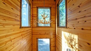 A wooden cabin interior with a central chandelier, large windows, and a door at the end of a narrow hallway.