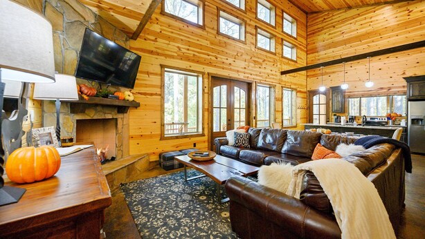 A log cabin interior with a stone fireplace, a leather sofa, a dining table, and a kitchen area.