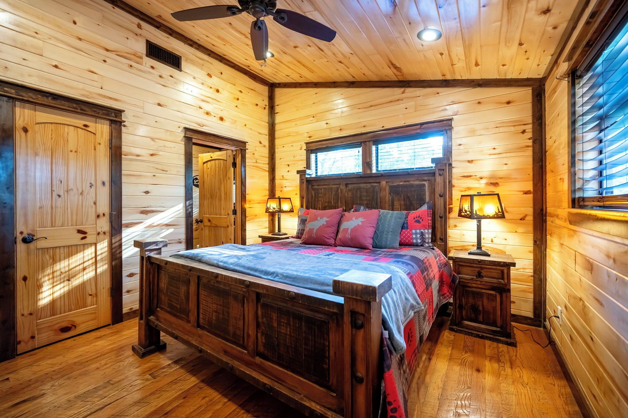 Ein Schlafzimmer einer Holzhütte mit einem Bett, zwei Nachttischen, einem Deckenventilator und einem Fenster mit Jalousien.