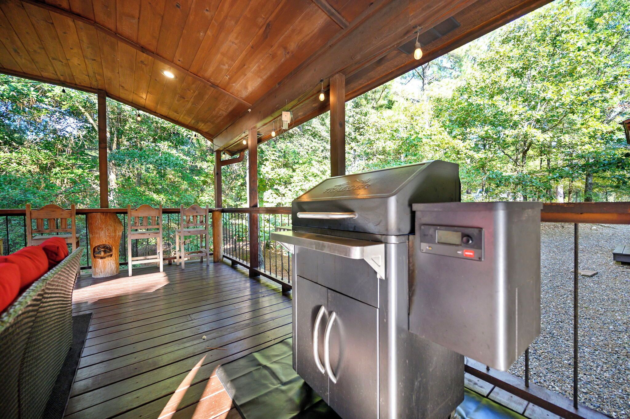 A covered outdoor area with a barbecue grill, wooden deck, and a bench with red cushions.