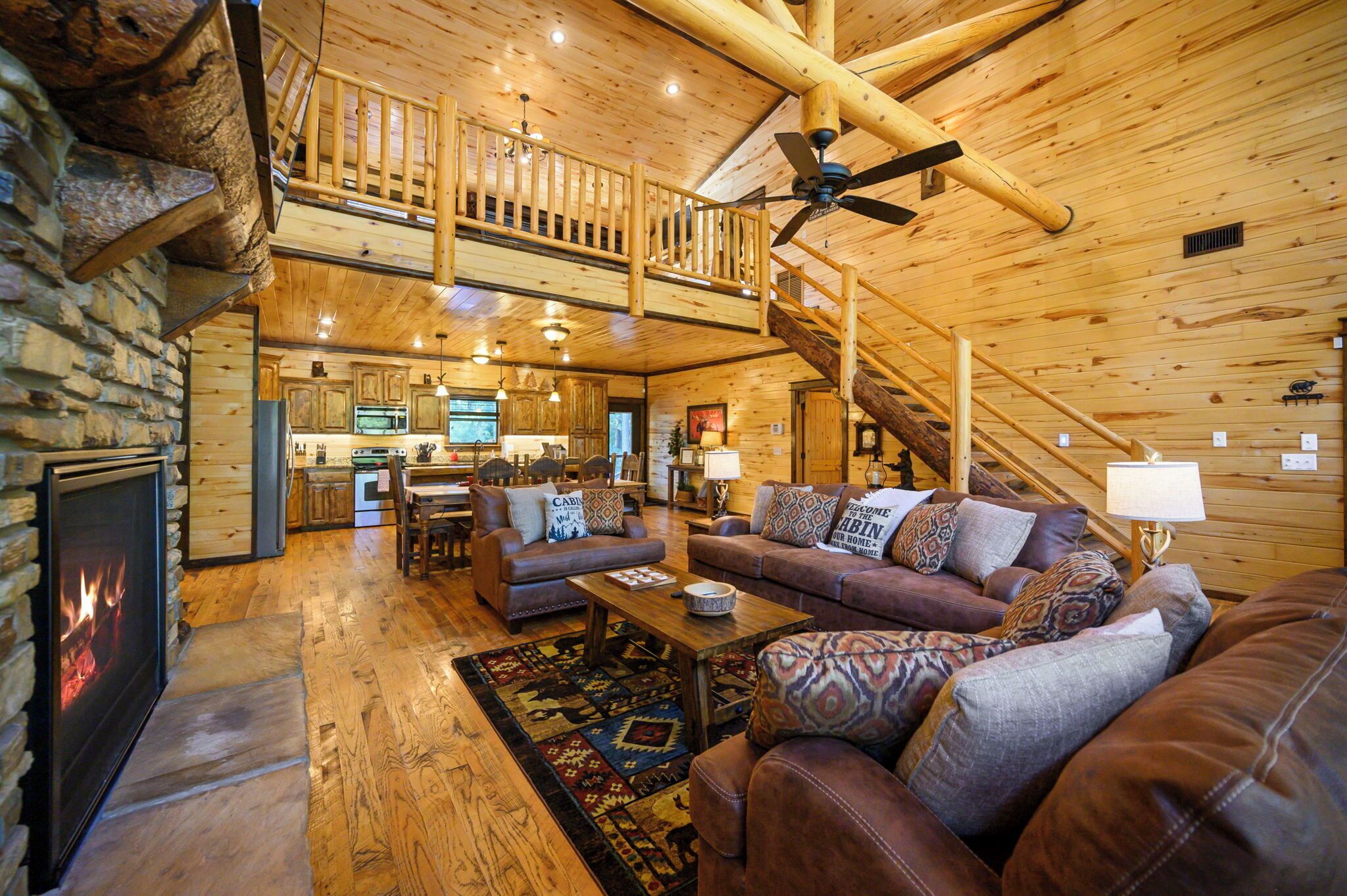 A spacious log cabin interior with a fireplace, a dining area, a living room with a sofa and two couches, and a staircase leading to an upper level.