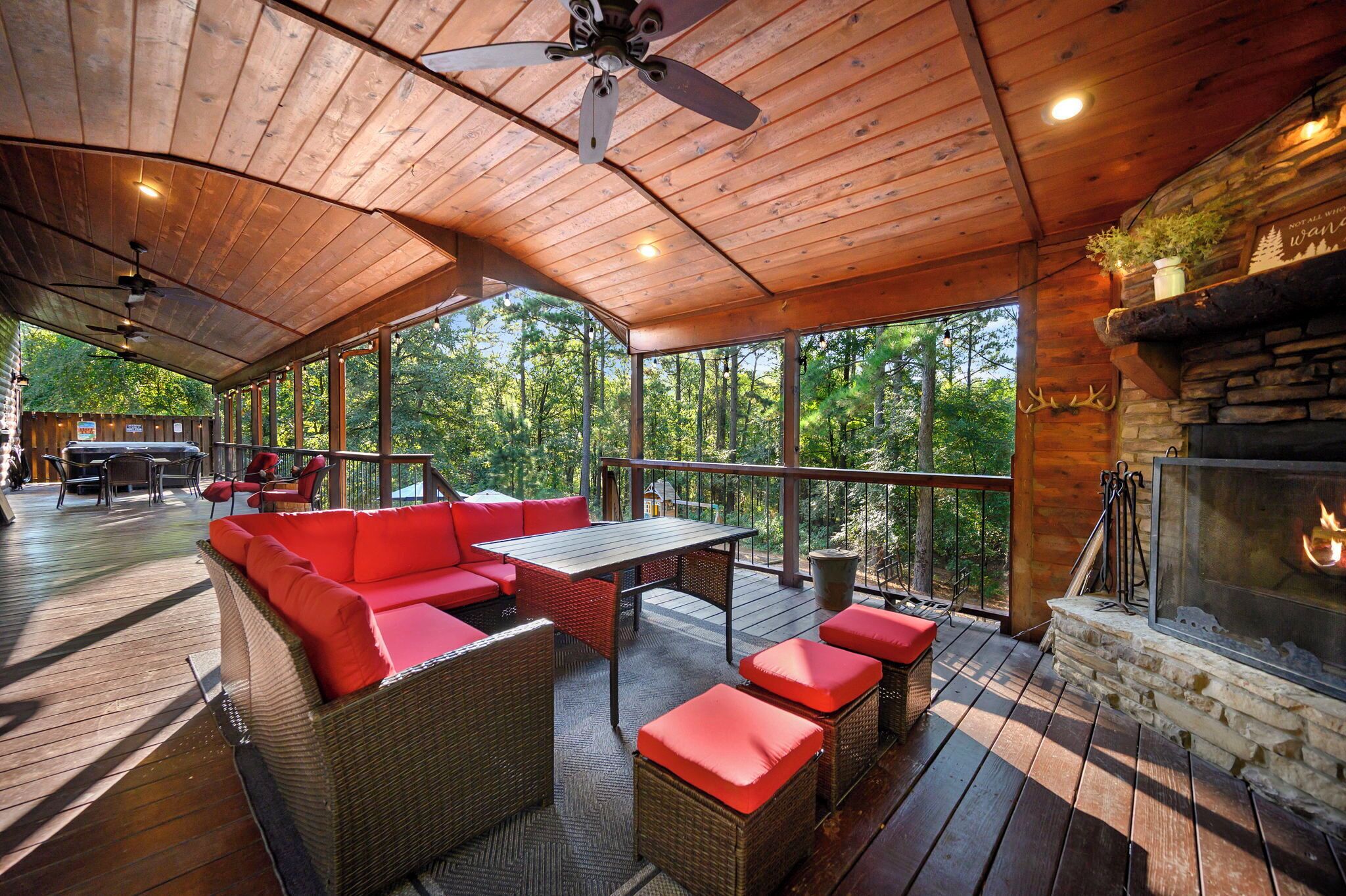 Un espace extérieur abrité, doté d’un plafond en bois, de meubles en osier avec des coussins rouges, d’une cheminée en pierre et d’une vue sur la forêt.