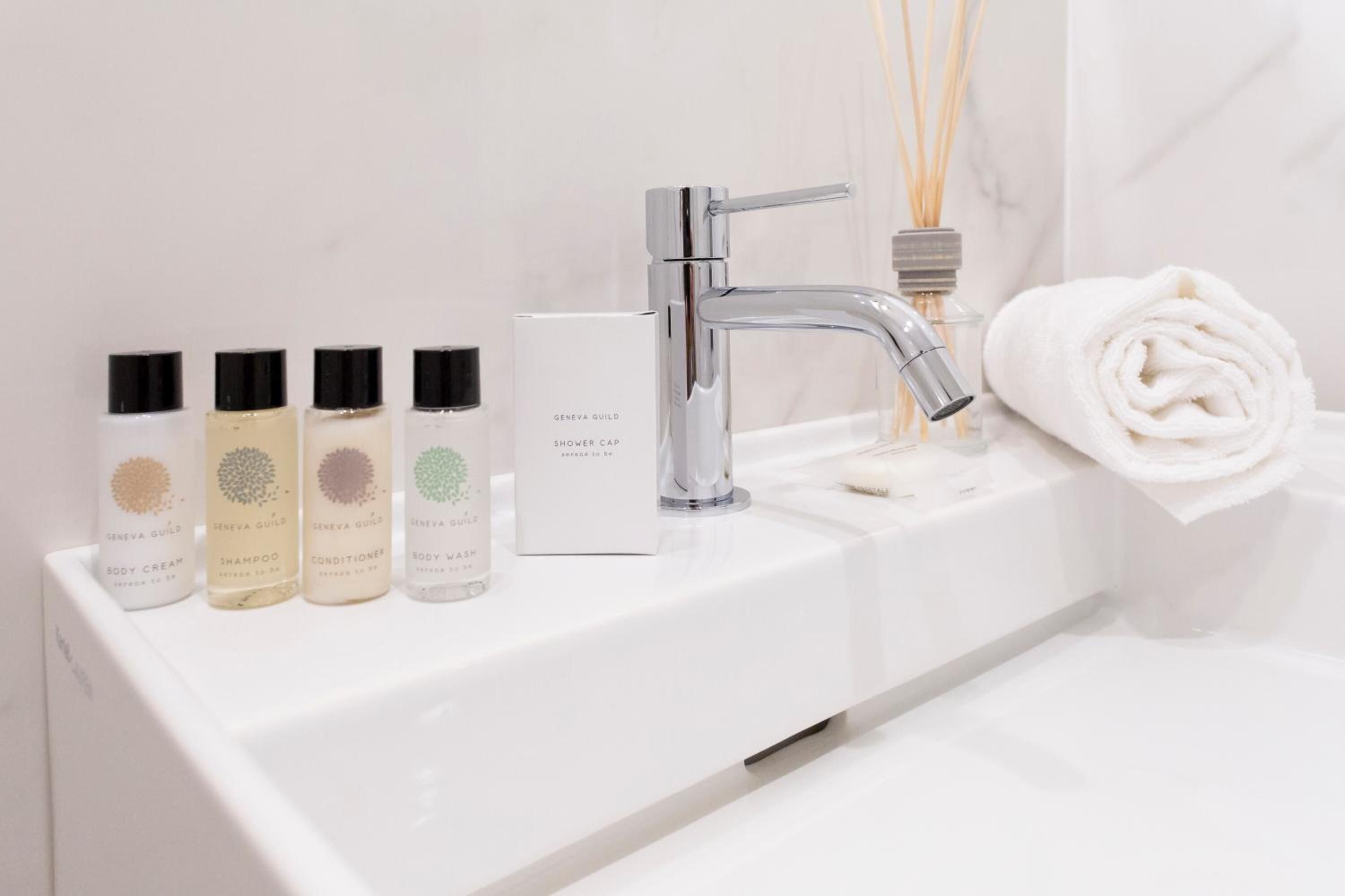 Un baño con un lavamanos blanco, un grifo cromado y cuatro botellas de productos para el cuidado corporal Geneva Guild.