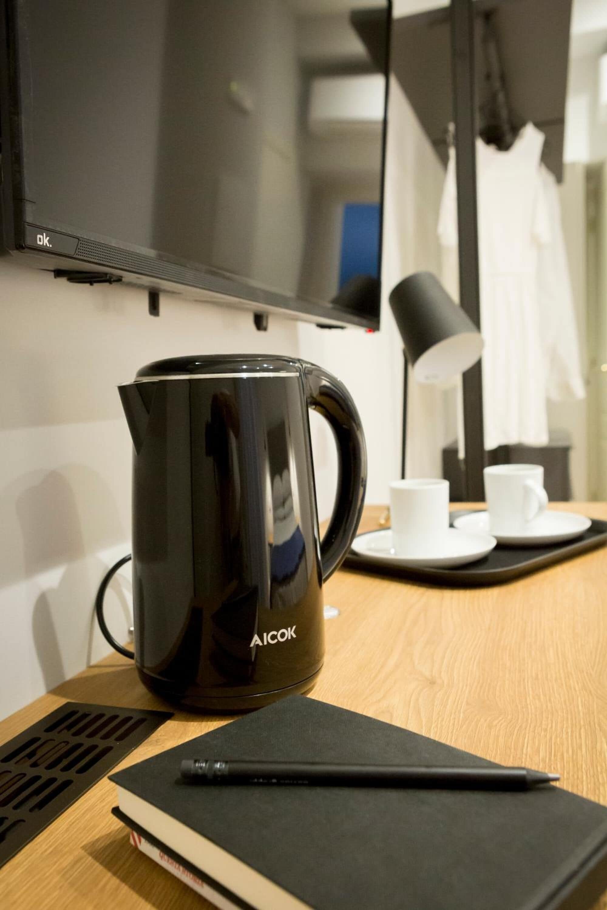 Una habitación de hotel moderna con una cafetera AICOK negra, un cuaderno y un bolígrafo sobre una mesa de madera.