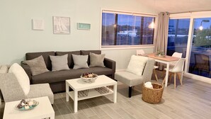 Una sala moderna con un sofá gris, una mesa de centro blanca y un sillón gris. Cuenta con una mesa de comedor con sillas, una ventana con persianas y un ventilador de techo.