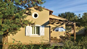 Einbungalow mit rundem Fenster, einer überdachten Terrasse mit Tisch und Stühlen sowie einem kleinen Garten.