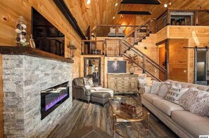 Cozy cabin living area with a stone fireplace, comfortable seating, and a staircase leading to additional living space.