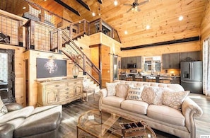 A spacious living area with a high ceiling, wooden beams, a staircase, a kitchen, and a dining area.