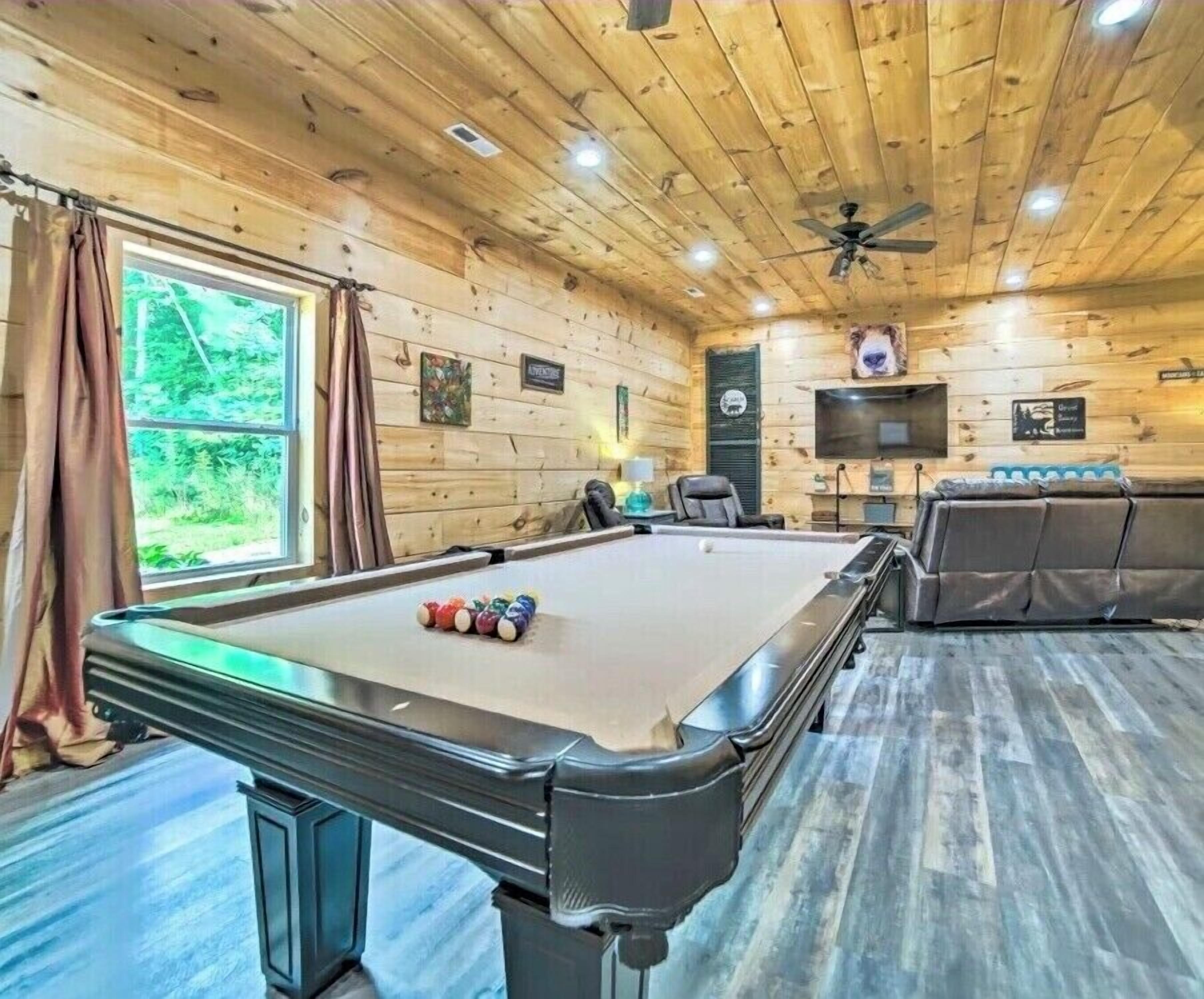 A pool table with balls, a sofa, a television, and a ceiling fan in a room with wooden walls and a window.