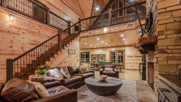 A spacious log cabin interior with a central seating area, a staircase, and a stone fireplace. - Boomerang Cabin | Large Family Retreat Near Broken Bow Lake (Broken Bow)