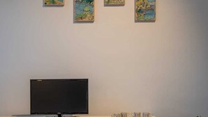Ein modernes Wohnzimmer mit einem Flachbildfernseher auf einer weißen Konsole, drei gerahmten Kunstwerken an der Wand und einer Zimmerpflanze rechts.
