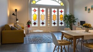 Ein Wohnzimmer mit einem gelben Sofa, einem Esstisch mit Stühlen, einem gemusterten Teppich und einem großen Fenster mit Buntglas.