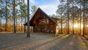A log cabin with a large front porch and multiple windows, surrounded by tall pine trees. - Bryce’s Crossroad | Family Cabin Near Broken Bow Lake (Broken Bow)